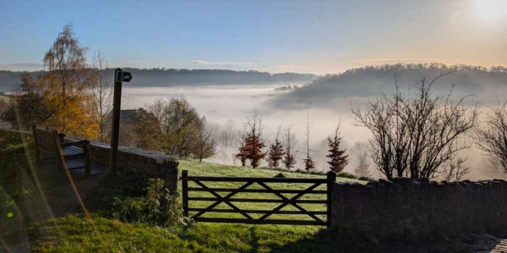 A wintery landscape showing a stone wall, a footpath sign and a gate. There are trees and hills in distance shrouded in morning mist,