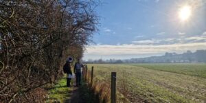 Two figures stand on a path next to a hedge. The spire of Salisbury cathedral can just about be seen in the distance. It's winter but the sun is shining.
