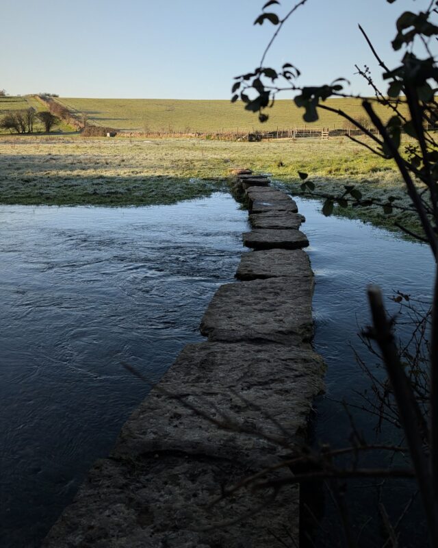 A view of stepping stones over a river with a green field stretching off into the distance.