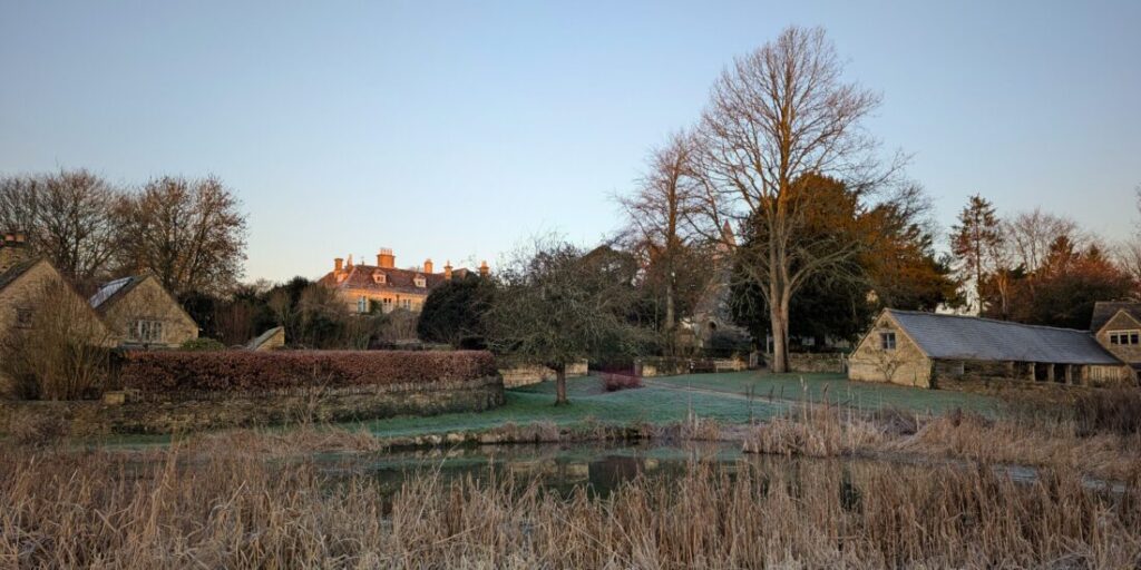 A Cotswolds village on a frosty morning, A village pond surrounded by reeds in the foreground with traditional Cotswolds stone buildings in the background.