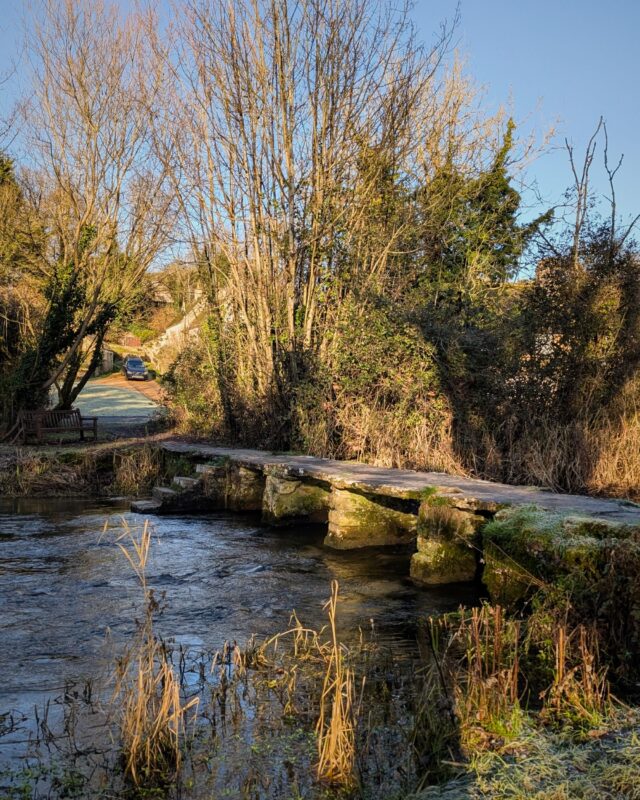 An old stone clapper bridge crossing a river in a wintery landscape. The golden sun is low in the sky, casting long shadows,