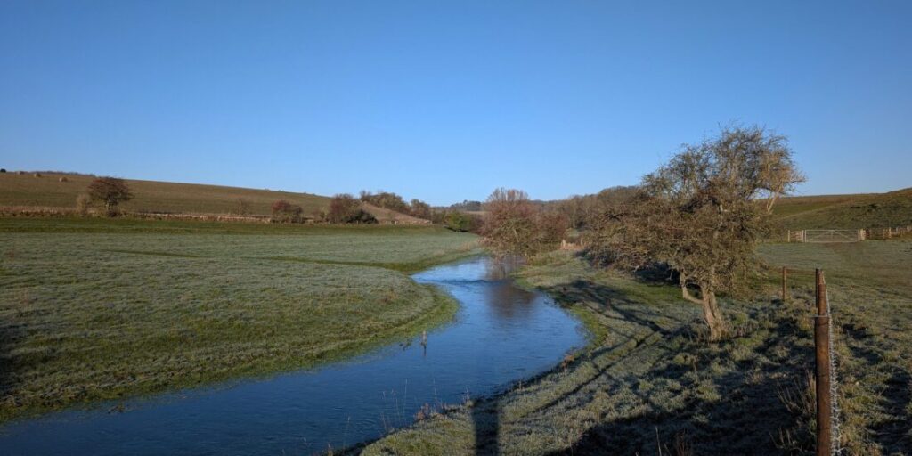 A river meanders through green countryside. The green grass has a covering of white frost.