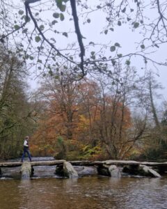 A person is walking across an ancient stone clapper bridge over a river surrounded by autumnal trees.