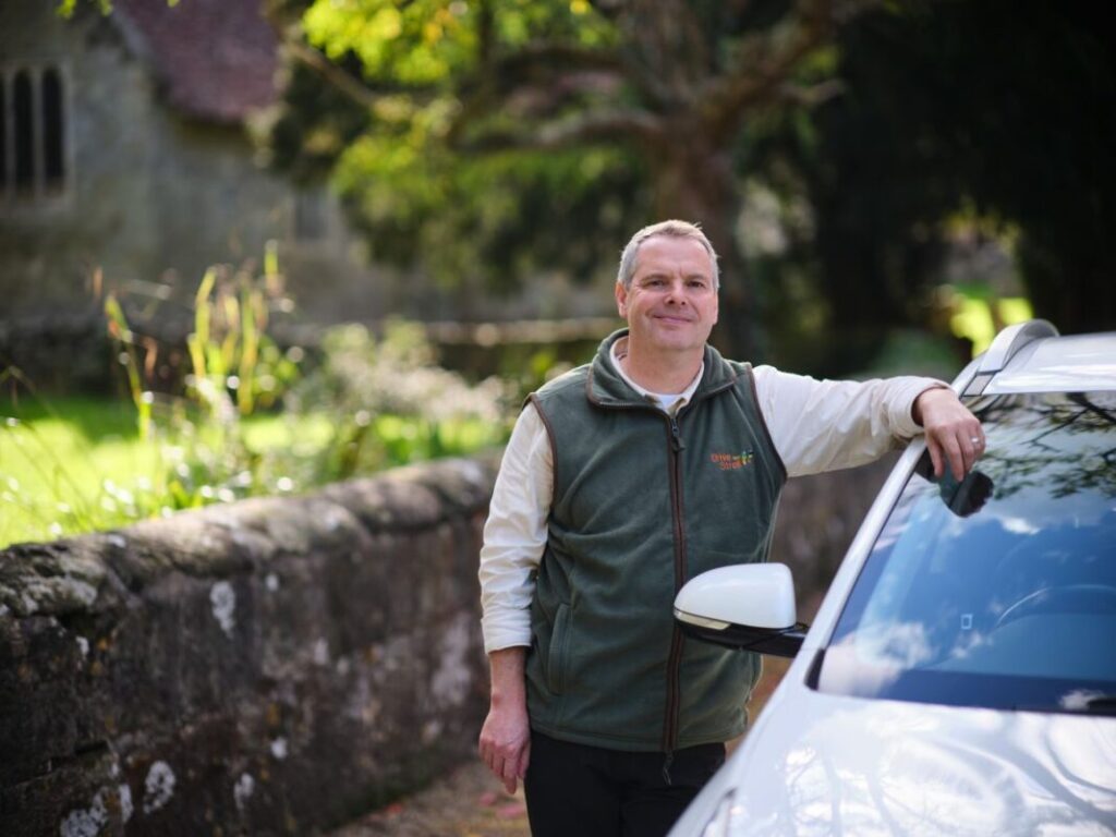 Foot Trails' Co-Founder David Howell in the countryside, standing next to a white car wearing a Drive and Stroll gilet.