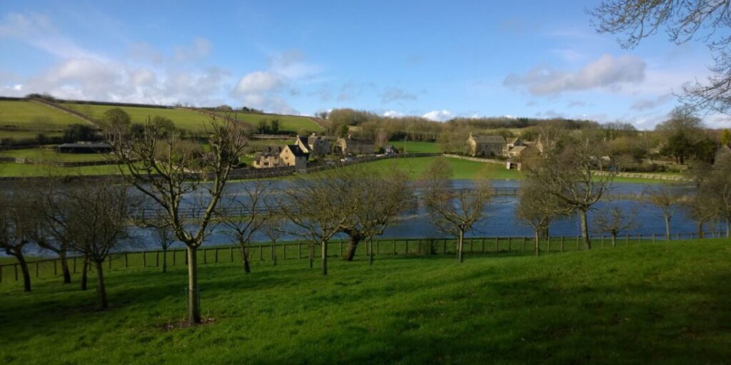 View across fields to a river and a collection of old stone houses in the Cotswolds