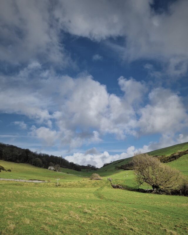 A dramatic valley with green hills on either side, trees in the distance and a footpath winding through the green fields.