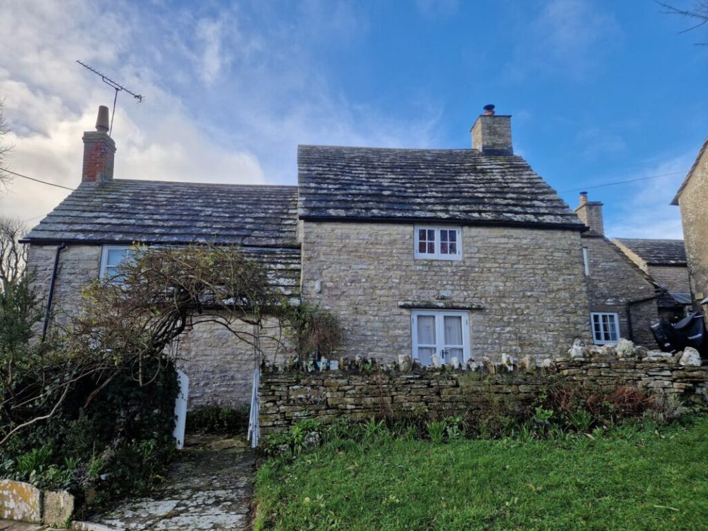 A grey stone cottage with a slate roof. There is a drystone wall with tree arching over the gate