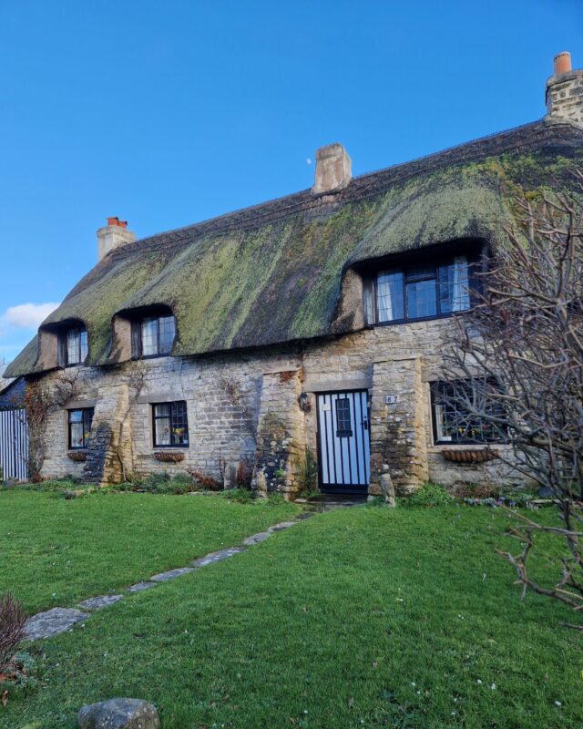 An old stone cottage with a thatched roof and a green lawn out front.
