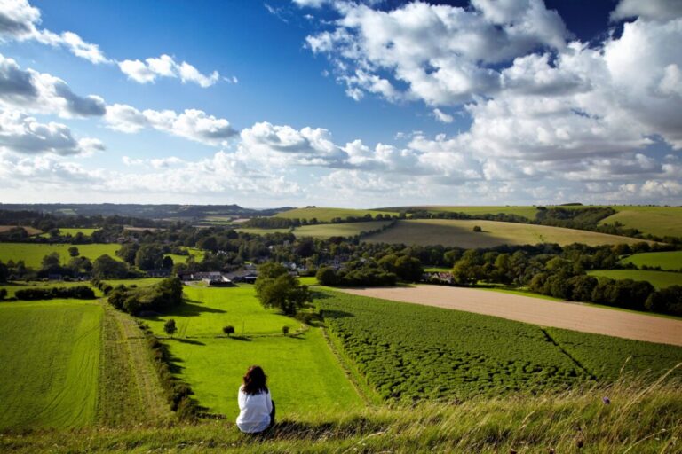 A female hiker is sat on the edge of a hill looking out over a patchwork of green farmland