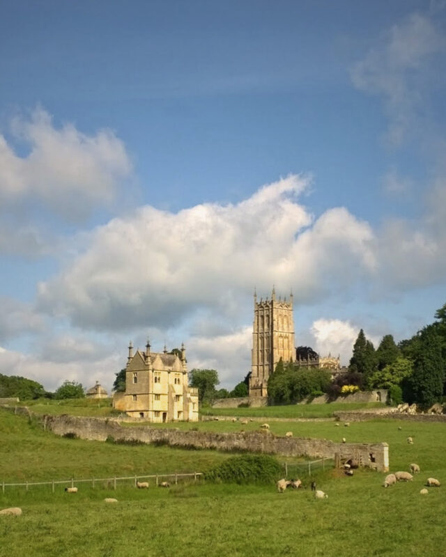 View of a classic Cotswolds wool church next to a grand country house. In the foreground are green fields and sheep.