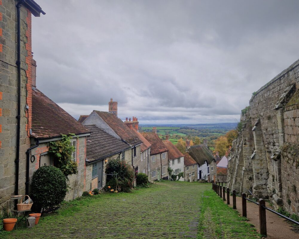 View looking down the ancient cobbled Gold Hill in Shaftesbury with thatched cottages on the left and the old castle wall to the right.
