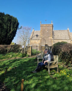 A walker sits on a bench in the spring sunshine in front of a 12th century church.