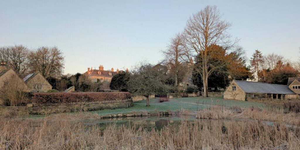 Image of a Cotswolds village on a frosty winter morning with a manor house, cottages & a barn set around a pond.