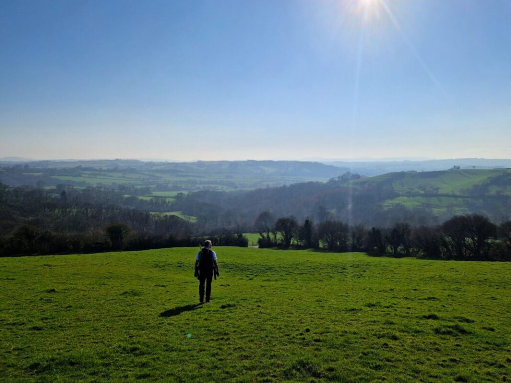 A lone walker strides down a hill towards a stunning view of fields and hills stretching off into the distance.