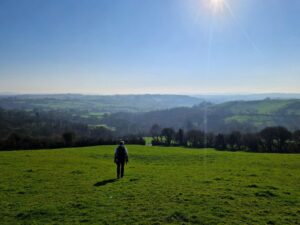 A lone walker strides down a hill towards a stunning view of fields and hills stretching off into the distance.