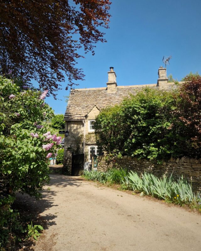 View of a lane at the end of which is a traditional stone Cotswolds cottage half hidden behind trees and hedges.