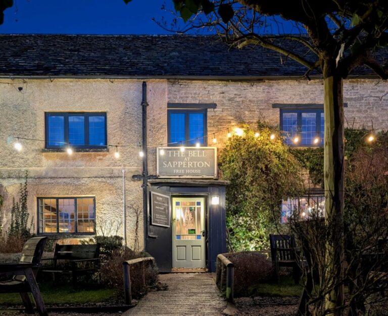 The front of an old stone country inn at night in the Cotswolds. A stone path leads to the front door and there are lights strung up outside glowing gently in the dark.
