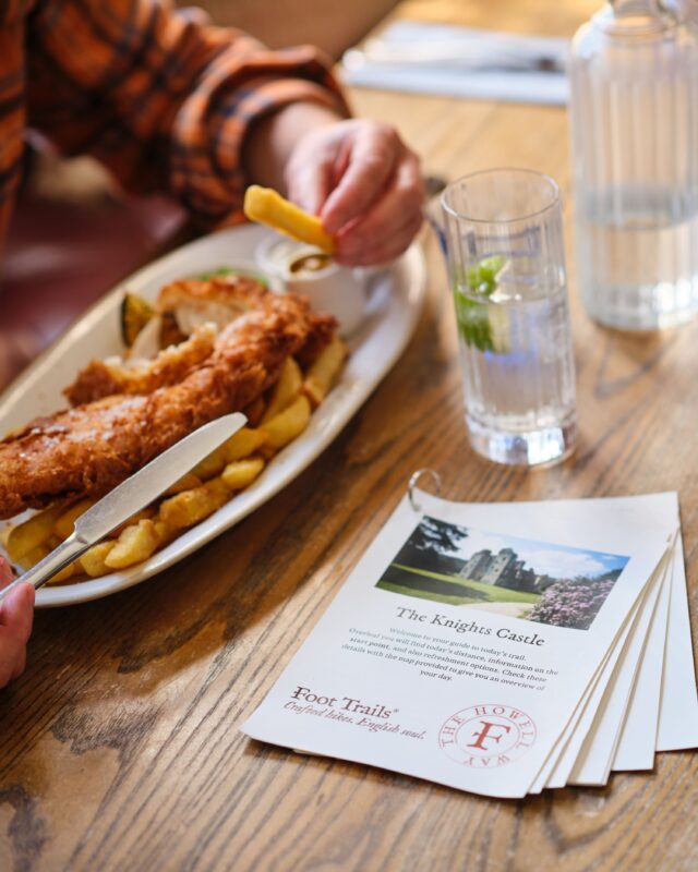 A person seated at a table with a plate of fish and chips. A glass of water and a Foot Trails trail guide are also on the table.