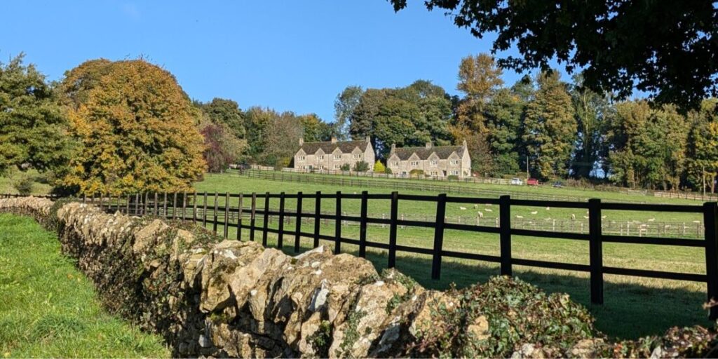 An autumn scene in the Cotswolds with a stone wall and a wooden fence running behind it. The leaves on the surrounding trees red and brown. In the background there is a row of cottages.