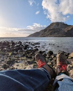 View of a hiker's legs and boots resting on the shore of a secluded cove with the sea and cliffs in the background.