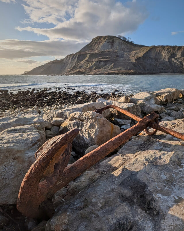 An old rusty anchor lies on a rocky shoreline with the sea and an imposing cliff in the distance.