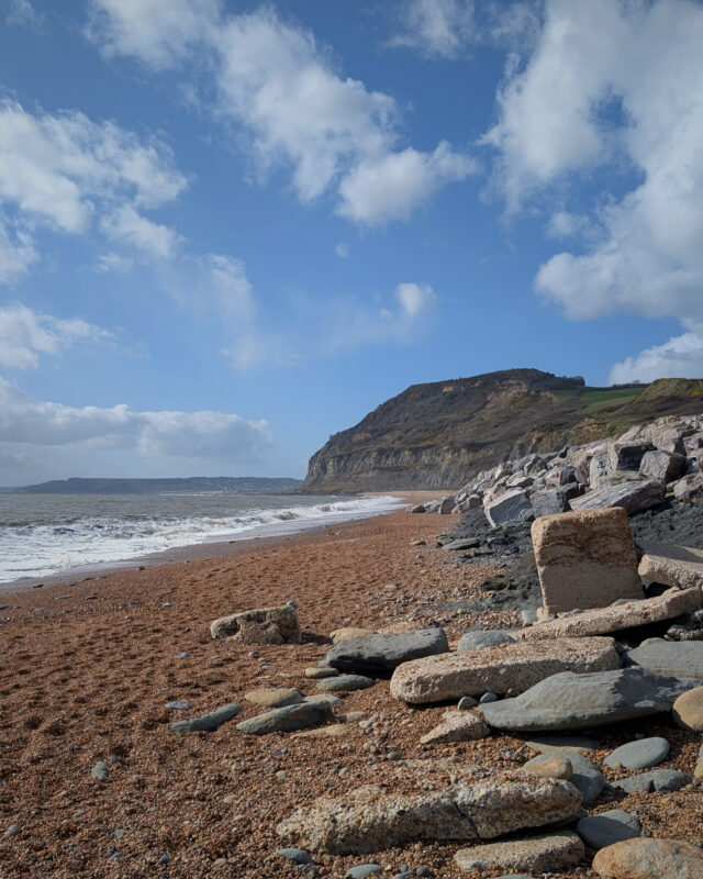 View of sea and cliffs in the distance from a shingle beach. The sun is shining and there are white clouds in the sky