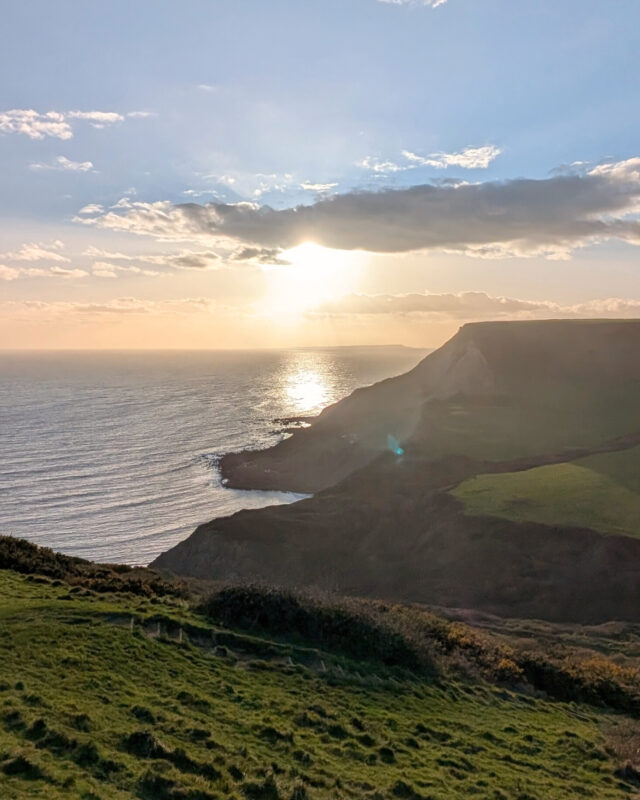 View of the coast from a cliff top with the sun low in the sky, just above the sea.
