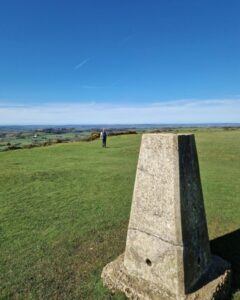 View of a trig point in the foreground with a views over the countryside. In the distance a hiker is walking off to the other side of the hilltop.