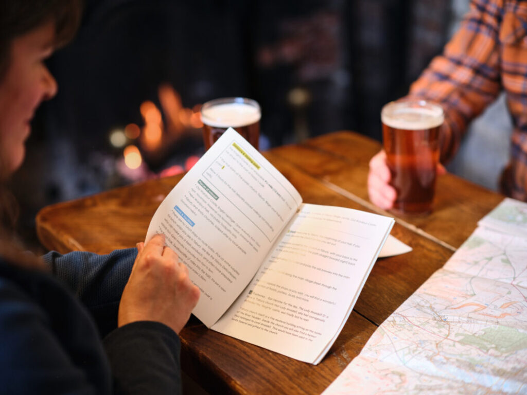Close up of two people's arms resting on a wooden table in a country inn with a wood fire in the background. One person is holding a pint of ale, the other is holding a trail guide. An OS map is laid on the table too.