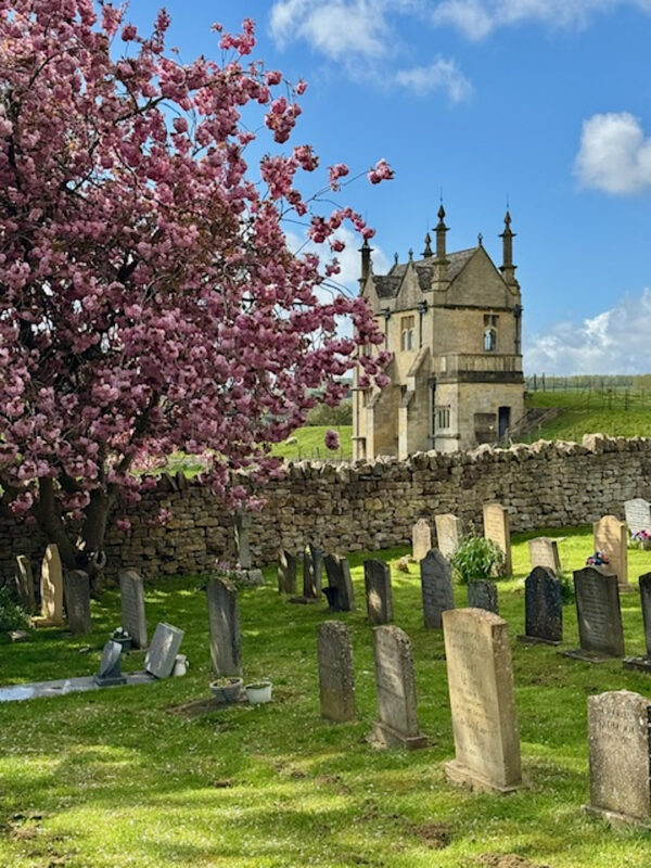 Photo of an ornate house in the Cotswolds taken from a churchyard. The house is behind an old church wall with tree blossoms and gravestones in the foreground.
