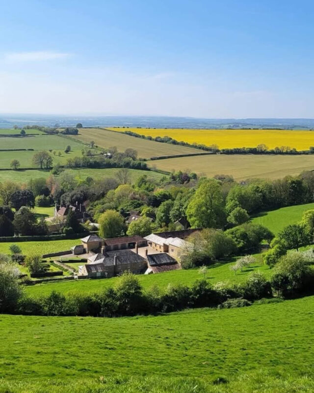 View of the English countryside from a hill, with the land sloping down to farm buildings and beyond, a patchwork of green farm fields