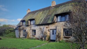 A thatched cottage with green moss growing on the thatch and a black and white striped front door leading out into a garden with a flagstone path