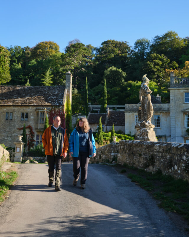 Two hikers are walking over an ornate stone bridge in front of a beautiful country mansion.