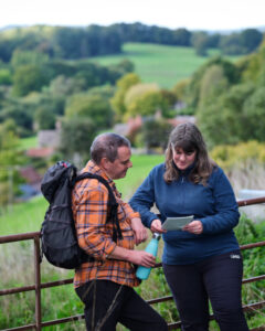 Two hikers are leaning against a fence, looking at a map. One of them is holding a water bottle.