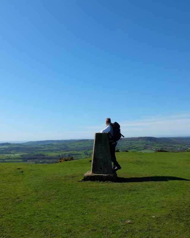 A hiker stands at a trig point on top of a hill against a bright blue, cloudless sky.