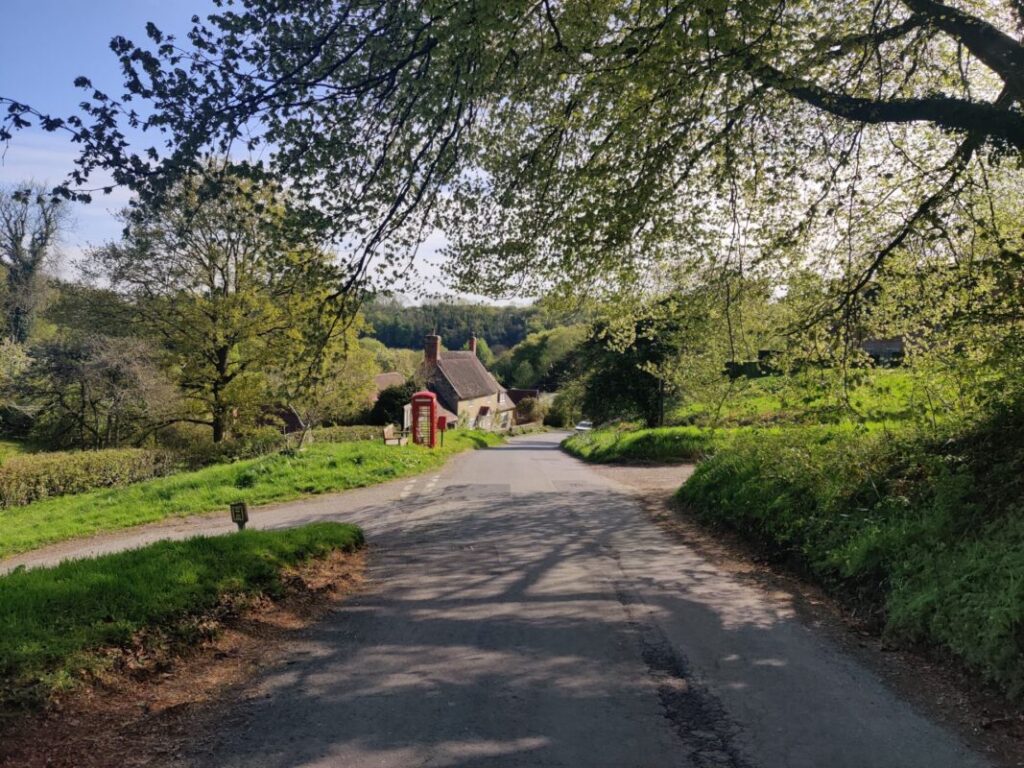 View of a quiet country lane with a red telephone box and cottage in the distance.