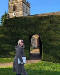 Foot Trails Co-Founder Alison standing in front of a large hedge with an old church tower appearing over the top of the hedge.