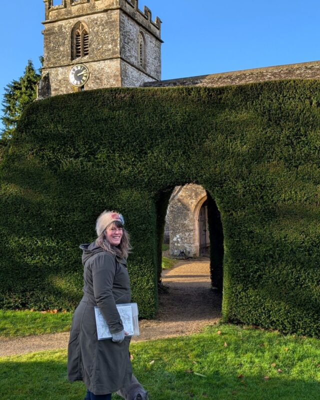 Foot Trails Co-Founder Alison standing in front of a large hedge with an old church tower appearing over the top of the hedge.