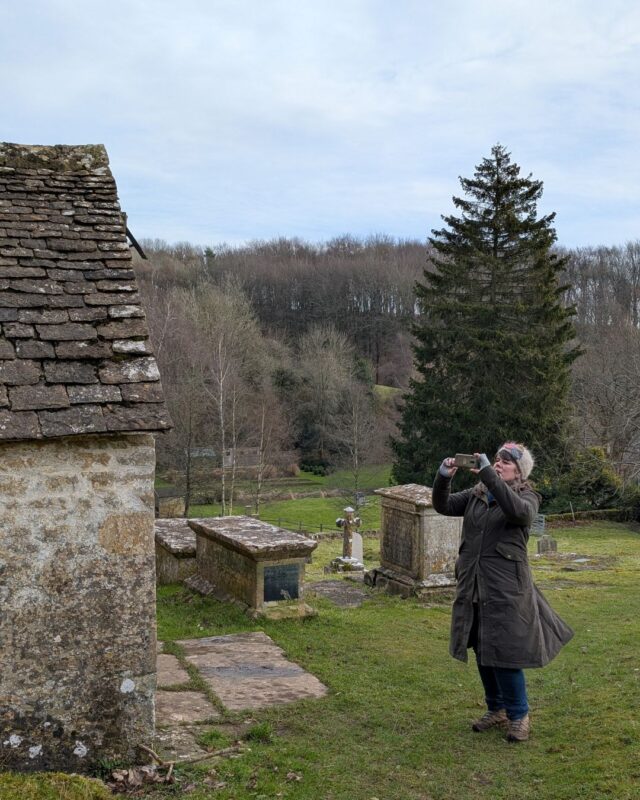 A female hiker is stood in a churchyard taking a photo of the church with her phone.