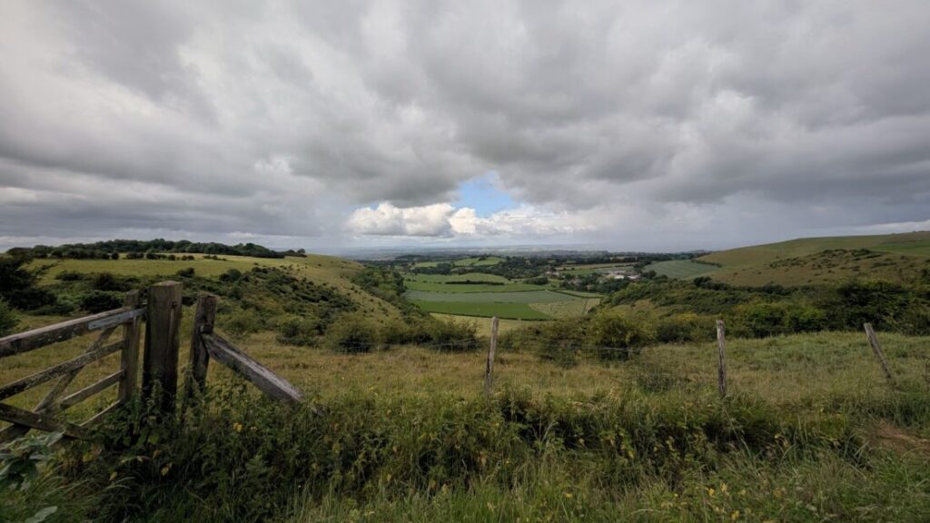 View over a valley of patchwork farm fields nestled between green hills with a wooden gate to the right