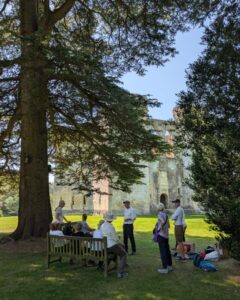 Guide David Howell stands with guests in the shade of trees explaining the history of Wardour Castle. The castle is visible in the background