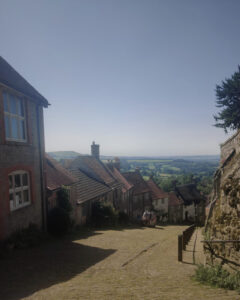 Photo of a steep cobbled street stretching off downhill. The cobbled street is flanked on the left by old cottages and by an old stone wall on the right. This is Gold Hill in Shaftesbury.