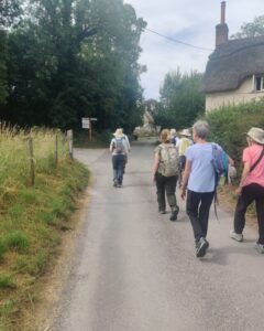 A group of hikers are walking down a lane in a village towards a junction with a thatched cottage to their right.
