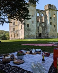 A white table cloth lies on the ground with plates and bowls of food on it. Wardour Castle ruins stand in the background.