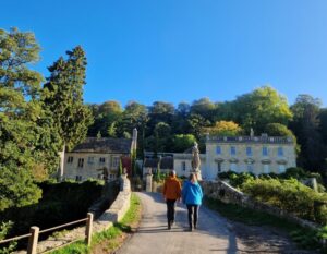 Two hikers shown from behind walking over an old stone bridge towards a manor house