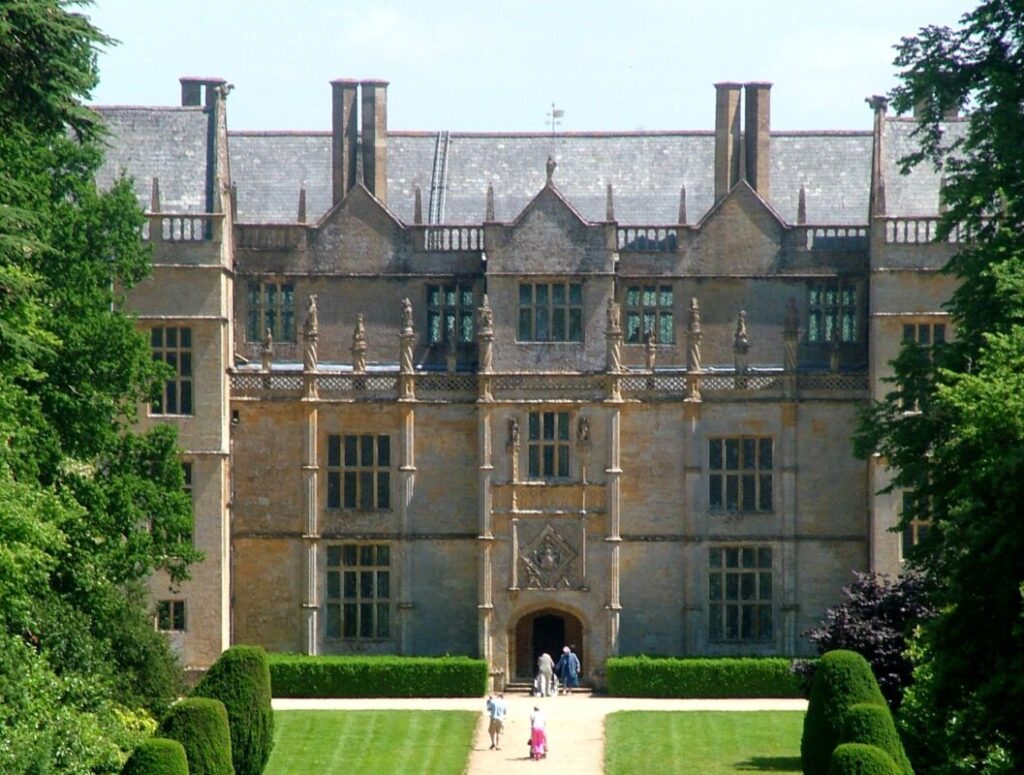 Front view of an Elizabethan manor house with people walking up a wide driveway through green lawns to the front door.
