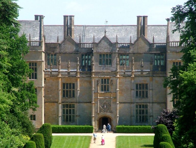 Front view of an Elizabethan manor house with people walking up a wide driveway through green lawns to the front door.