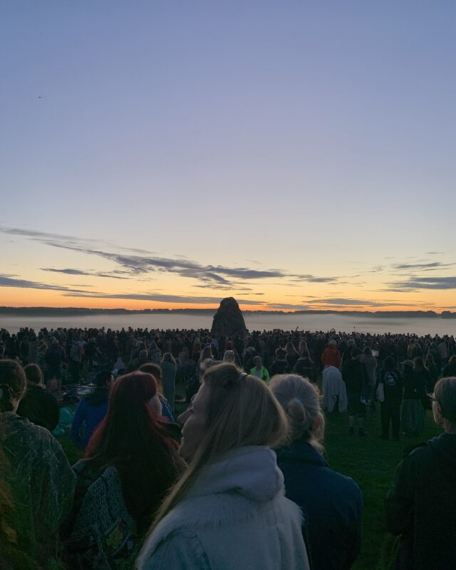 View of the sun rising over crowds and a standing stone at Stonehenge.