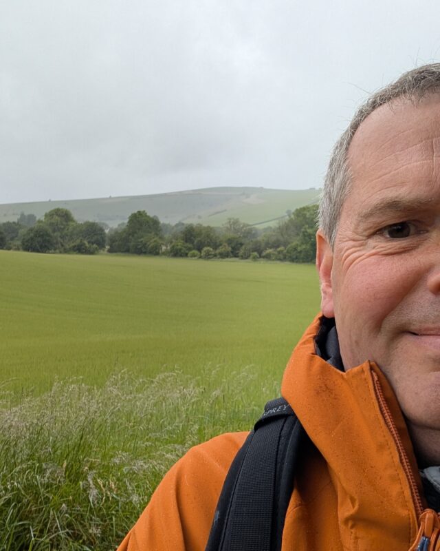 A selfie taken by Foot Trails Co-Founder David showing a field, trees and a hill behind him under a grey, cloudy sky.
