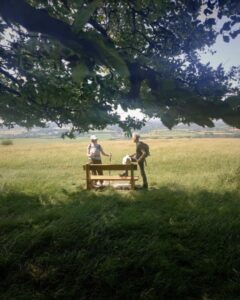 Two hikers standing next to a bench and talking. They are on top of a hill with fields and hills stretching off into the distance.